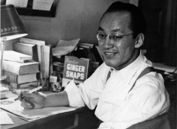 Black and white photograph of John Okada, sitting at a desk covered in books and holding a pen.